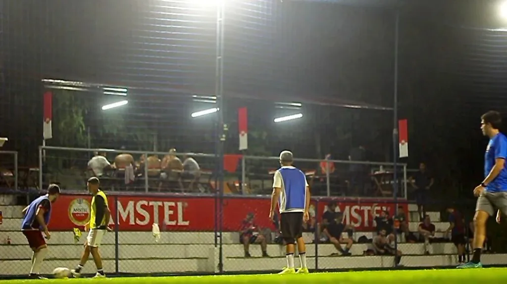 Jogo de futebol society à noite na Arena Society Show, no Leblon, com iluminação profissional e arquibancada ao fundo